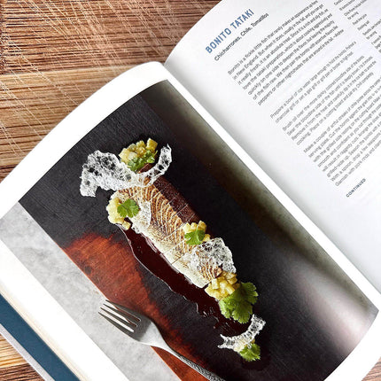 Gourmet dish on a dark surface with a fork, open cookbook displaying text and images.
