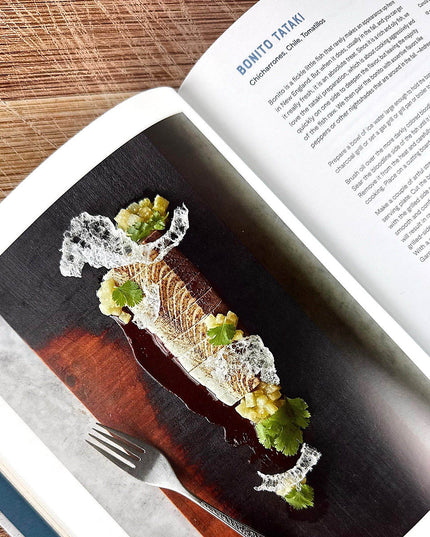 Gourmet dish on a dark surface with a fork, open cookbook displaying text and images.