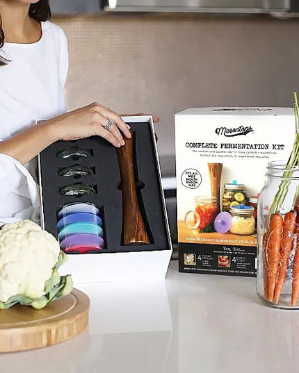 a model at a countertop holding the opened package of the mason jar fermentation tool kit with raw cabbage on the counter along with a bunch of carrots in a mason jar