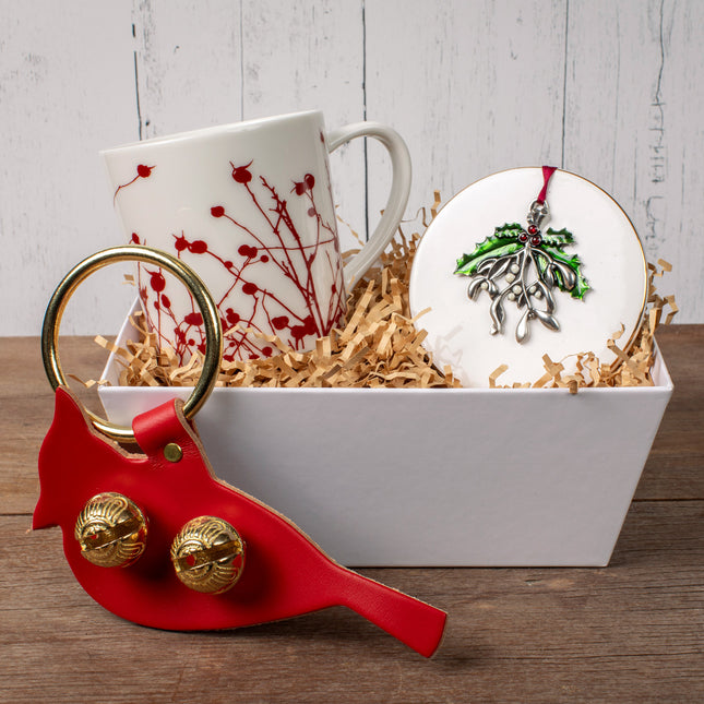 the festive accents gift box on a wood table - the winterberries mug and mistletoe ornament are arranged in a white tray surrounded by kraft colored crinkle packing and the cardinal door hanger bell sits in front