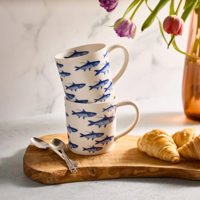 Two stacked ceramic mugs with blue fish pattern on a wooden board with croissants and spoons.