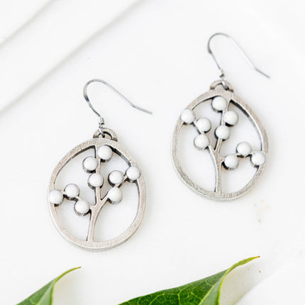 a pair of oval pewter earrings with branches inside adorned with white beads to represent blossoms on a white table next to some bright green leaves