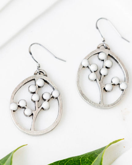 a pair of oval pewter earrings with branches inside adorned with white beads to represent blossoms on a white table next to some bright green leaves