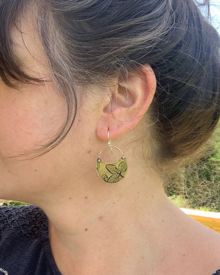 Close-up of a person wearing the hydrangea crescent earrings in brass with a natural background