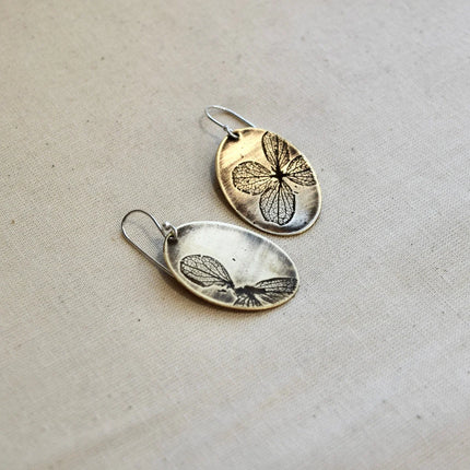 Pair of oval shaped brass earrings with pressed hydrangea petals designs on a beige background
