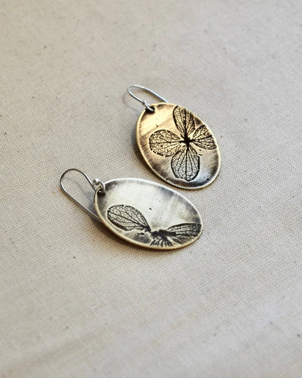 Pair of oval shaped brass earrings with pressed hydrangea petals designs on a beige background