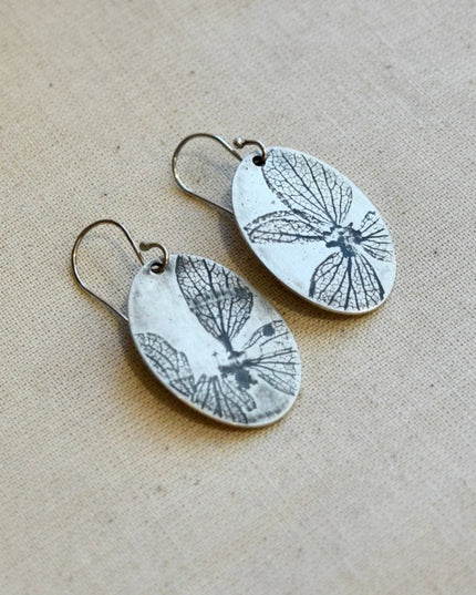 Pair of oval shaped silver earrings with pressed hydrangea petals designs on a beige background