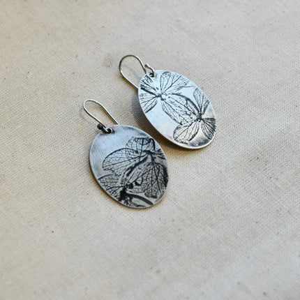Pair of oval shaped silver earrings with pressed hydrangea petals designs on a beige background