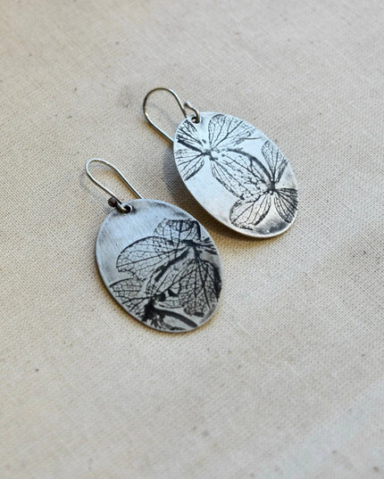 Pair of oval shaped silver earrings with pressed hydrangea petals designs on a beige background