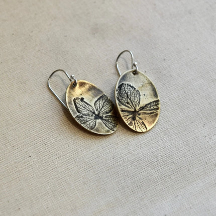 Pair of oval shaped brass earrings with pressed hydrangea petals designs on a beige background