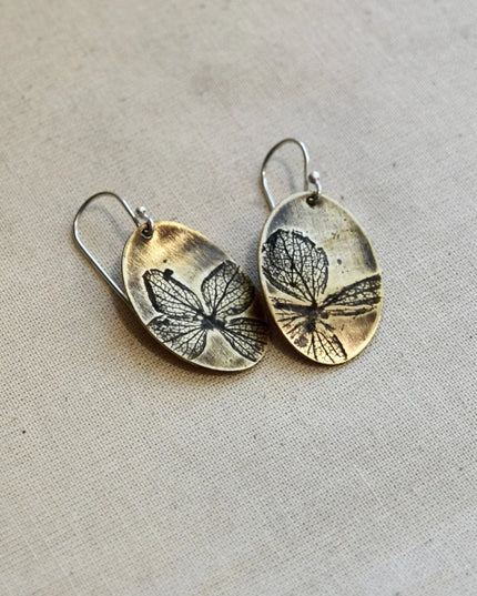 Pair of oval shaped brass earrings with pressed hydrangea petals designs on a beige background