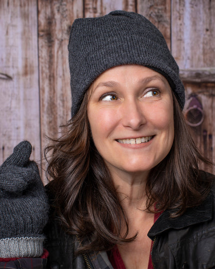 a smiling woman stands in front of a rustic barn door and wears the gray iditarod hat and matching gloves