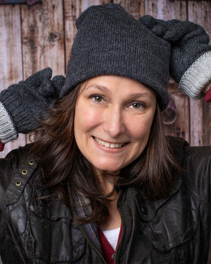a smiling woman stands in front of a rustic barn door wearing the gray iditarod hat and matching gloves - she holds her hands behind her head playfully