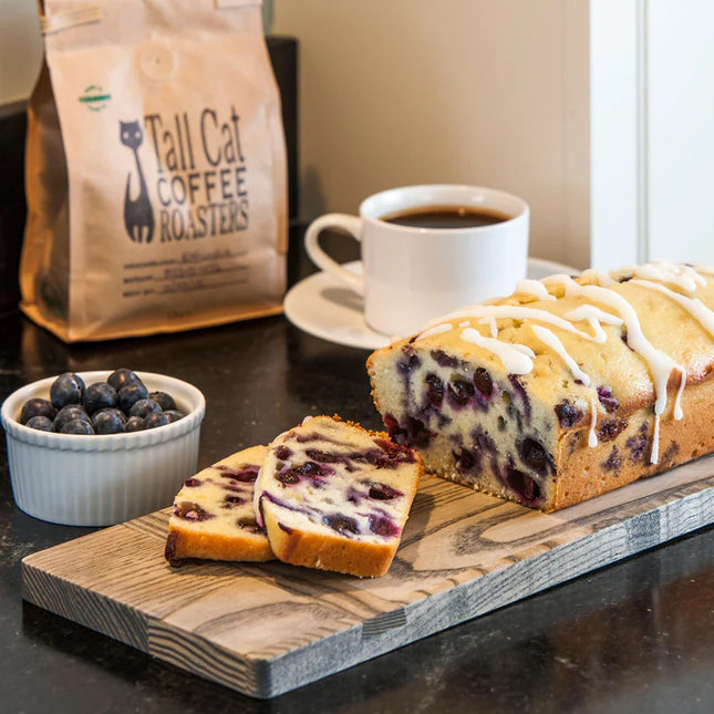 Blueberry cake with white icing on a wooden board, next to a cup of coffee and a bag of Tall Cat Coffee Roasters.