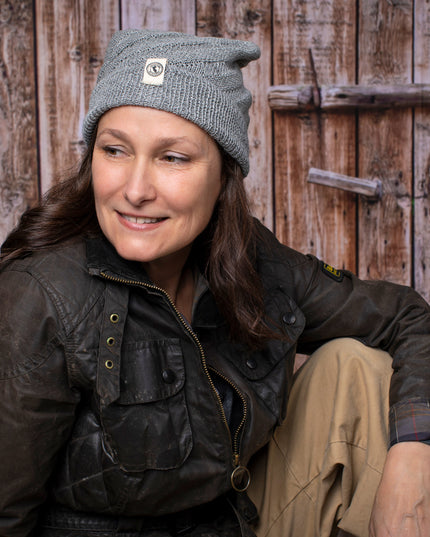 a fair skinned, brunette female model wearing the Kingsley beanie in Gray. She is smiling and posing casually with her arm resting on her knee and sitting in front of a rustic barn door