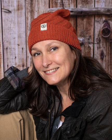a fair skinned, brunette female model wearing the Kingsley beanie in Rust. She is smiling and posing casually with her arm resting on her knee and sitting in front of a rustic barn door