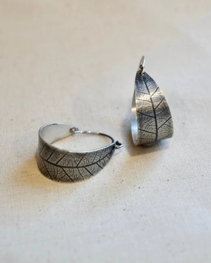 Silver leaf-patterned hoop earring and pendant on a beige fabric background