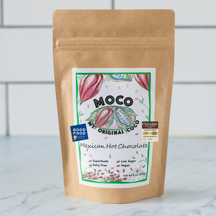 a kraft hot cocoa package with a white label with red and green illustrations of cocoa beans that reads "MOCO My Original Coco - Mexican Hot Chocolate"
