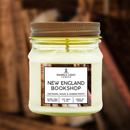 a candle in a clear glass mason jar with off white wax and a label that reads "New England Bookshop" against a soft image of a bookcase