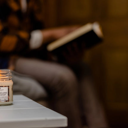 a New England Bookshop candle burning on a side table next to someone seated and reading