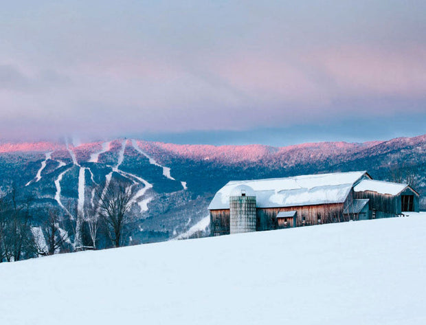 Snow-covered landscape with a barn and ski slopes under a pink and blue sky.