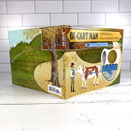 Book titled 'Ox-Cart Man' by Donald Hall on a white wooden surface with a white tiled wall background.