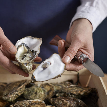 Person opening an oyster with a knife over a tray of oysters.
