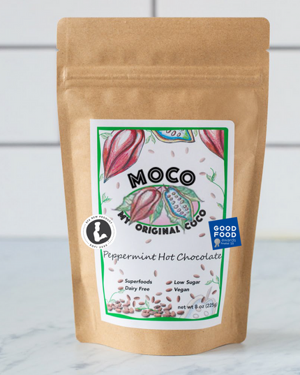 a kraft hot cocoa package with a white label with red and green illustrations of cocoa beans that reads "MOCO My Original Coco - Peppermint Hot Chocolate" on a marble counter with white subway tile background