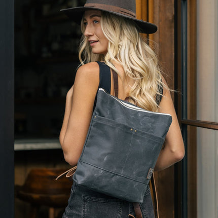 a blonde woman with a wide brim hat stands in a doorway with the Day Pack in Ash slung over her left shoulder