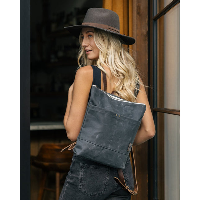 a blonde woman with a wide brim hat stands in a doorway with the Day Pack in Ash slung over her left shoulder
