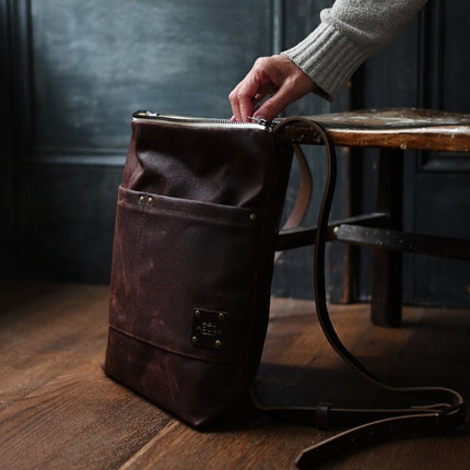 the day pack in chestnut rests against a chair while a hand gently reaches into the top of the pack