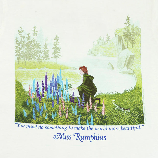 closeup of the tee shirt graphic showing Miss Rumphuis kneeling down to admire Lupine flowers against a coastal Maine landscape
