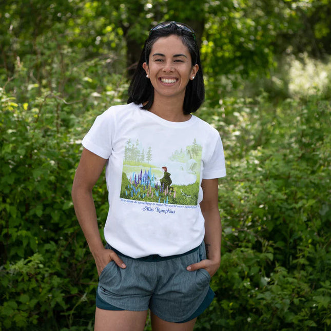 Woman wearing a white t-shirt with a Miss Rumphis illustration, standing in a green outdoor setting