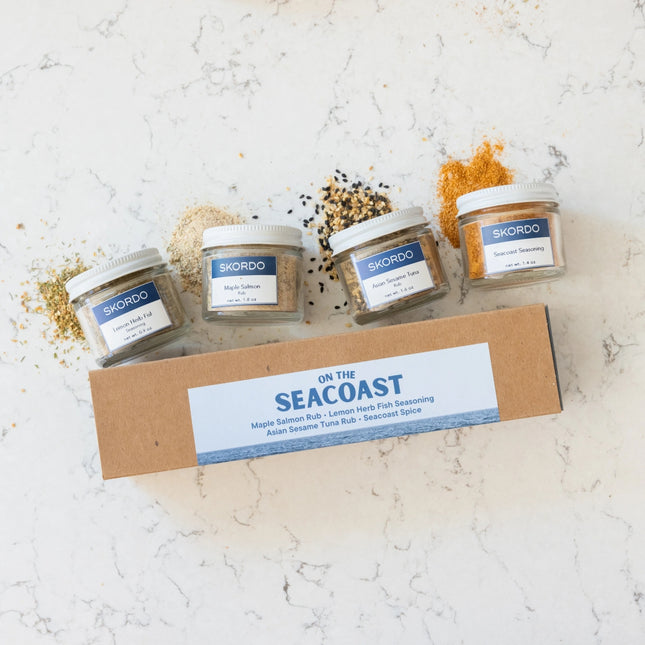 Four jars of Skordo seasoning on a cardboard box labeled 'On the Seacoast' on a light marble background.