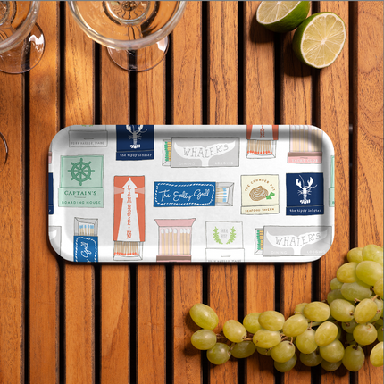 Decorative tray with a pattern of illustrated nautical themed matchbooks on a wooden surface with grapes and lime wedges.