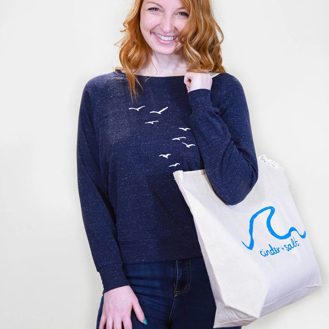 Woman wearing a navy blue sweater with bird designs, holding a white tote bag with a logo.