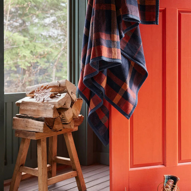 a rustic cabin scene with the sebago tartan blanket draped over an open, orange door with a pair of boots, chopped firewood, and a shearling rug nearby setting the mood for this cozy autumnal blanket