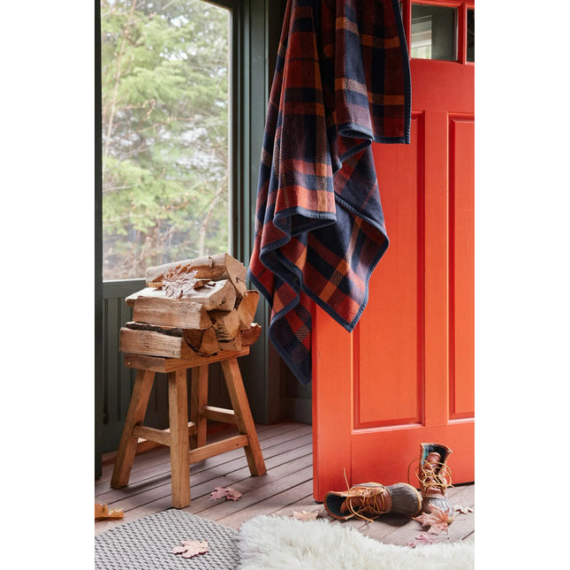 a rustic cabin scene with the sebago tartan blanket draped over an open, orange door with a pair of boots, chopped firewood, and a shearling rug nearby setting the mood for this cozy autumnal blanket
