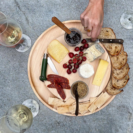 Wooden platter with assorted cheeses, meats, crackers, and bread on a concrete surface.