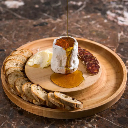 Wooden plate with a cheese wheel, bread slices, and honey being drizzled over the cheese.