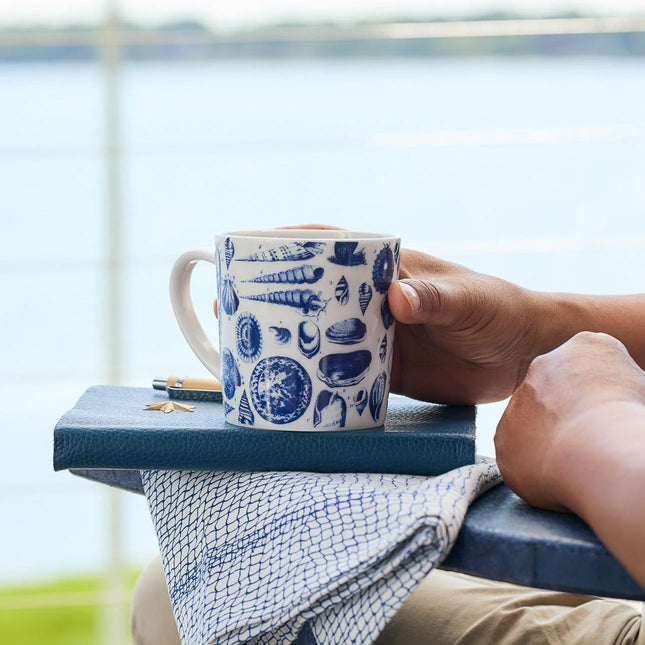Person holding a blue and white patterned mug with a book and pen on a surface by a window.