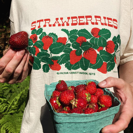 a close up of a model wearing the strawberries t-shirt while holding a quart of freshly picked strawberries