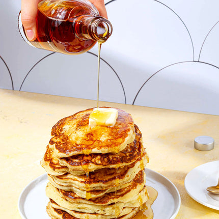 a bright kitchen table with a tall stack of pancakes on a plate with maple syrup being poured over them