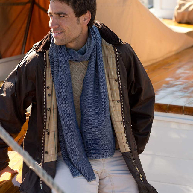Man wearing the indigo camden scarf and brown jacket standing on the deck of a sailboat