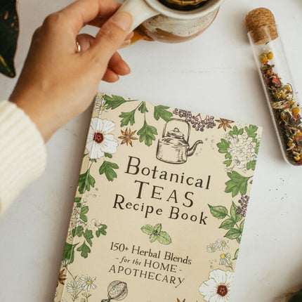 The botanical teas recipe book on a white table next to a vial of dried herbs next to a tea cup with strainer 