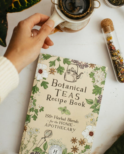 The botanical teas recipe book on a white table next to a vial of dried herbs next to a tea cup with strainer 