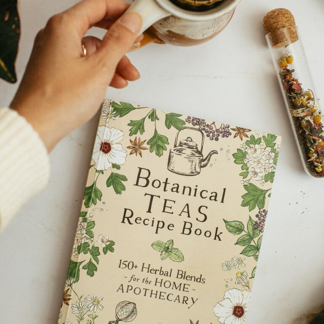 The botanical teas recipe book on a white table next to a vial of dried herbs next to a tea cup with strainer 