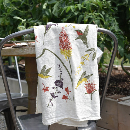 the hummingbird garden kitchen tea towel folded and layed over the back of a chair on a lush garden patio