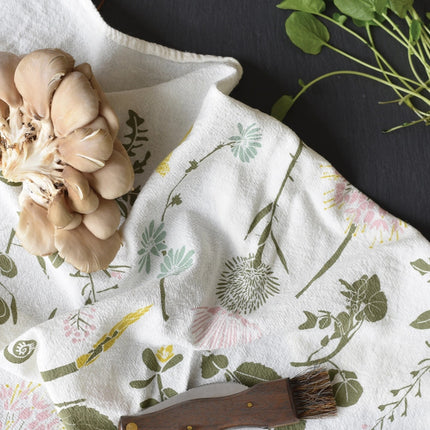 close up of the edible wilds tea towel in use holding foraged mushrooms, a foraging knife, and herbs