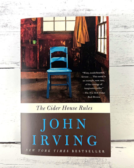 Book cover of 'The Cider House Rules' by John Irving with a blue chair in a room.